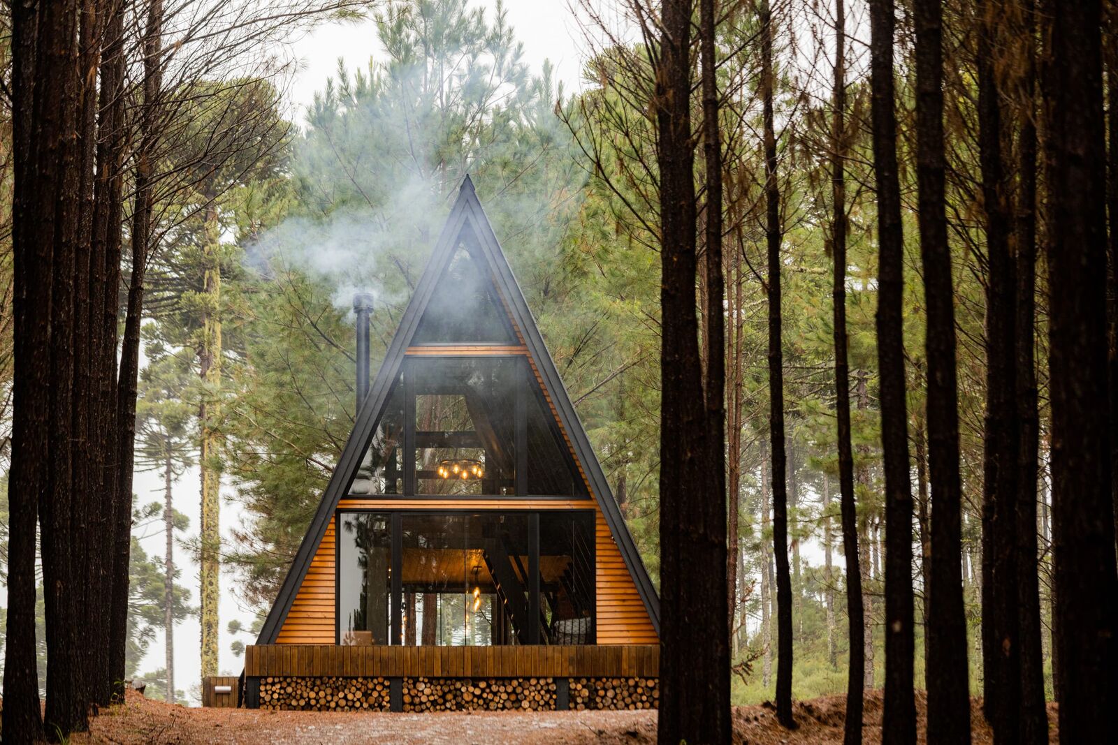Cabana A-frame de madeira escura com a lareira acesa e a fumaça subindo, emoldurada por troncos de pinheiros na névoa da Serra Gaúcha